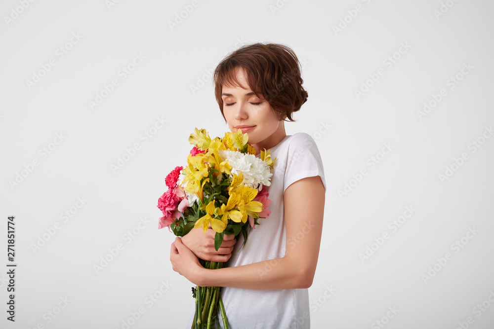 Young smiling nice short haired girl in white blank t-shirt, holding a bouquet of colorful flowers, enjoying the smell, standing over white background with closed eyes. Young smiling nice short haired girl in white blank t-shirt, holding a bouquet of colorful flowers, enjoying the smell, standing over white background with closed eyes.