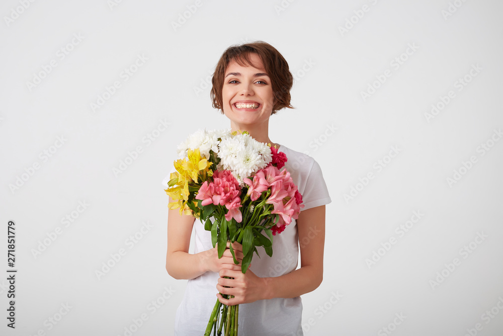Portrait of smiling nice short haired lady in white blank t-shirt, holding a bouquet of colorful flowers, very glad and smiling over white wall. Portrait of smiling nice short haired lady in white blank t-shirt, holding a bouquet of colorful flowers, very glad and smiling over white wall.