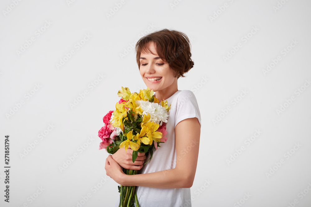 Cute happy young short haired girl in white blank t-shirt, holding a bouquet, bites his lip and looks at the of colorful flowers, enjoying the smell, standing over white background. Cute happy young short haired girl in white blank t-shirt, holding a bouquet, bites his lip and looks at the of colorful flowers, enjoying the smell, standing over white background.