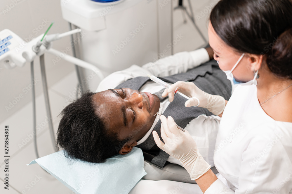 Obraz premium Young woman dentist treating root canals in the dental clinic. Young African American male with bad teeth lying on dentist chair with open mouth.