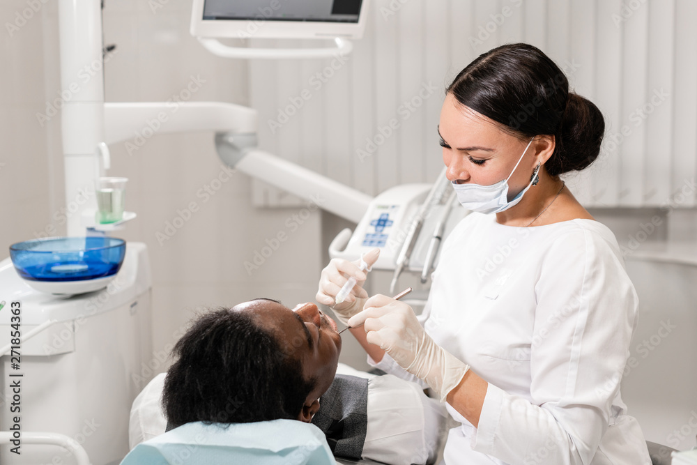 Young woman dentist treating root canals in the dental clinic. Young African American male with bad teeth lying on dentist chair with open mouth. Young woman dentist treating root canals in the dental clinic. Young African American male with bad teeth lying on dentist chair with open mouth.