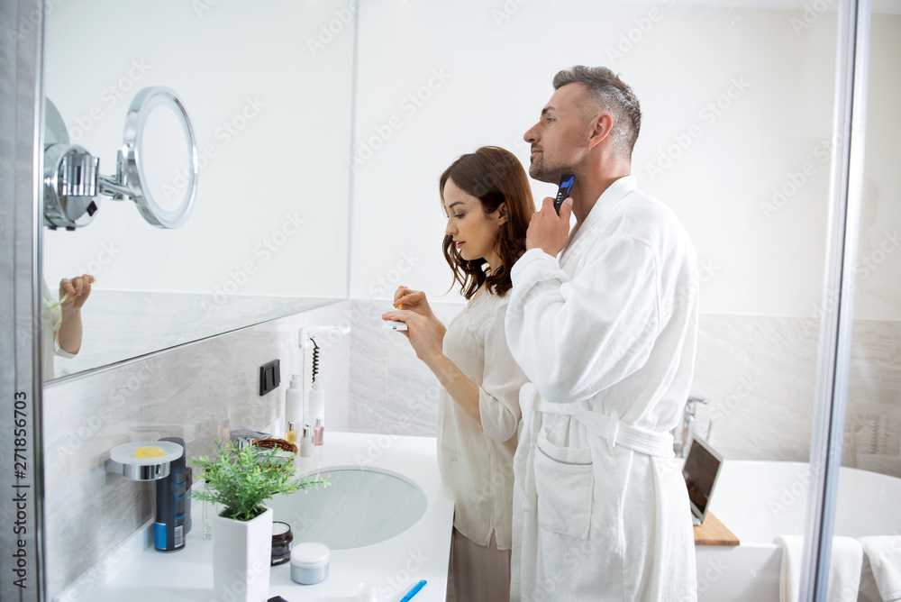 Fototapeta premium Woman brushing her teeth while smiling man shaving his beard
