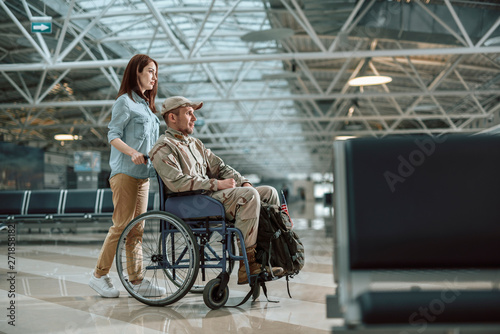 Beautiful wife carrying wheelchair and situating behind her husband in military uniform