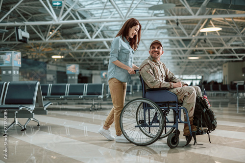 Beautiful wife carrying wheelchair and situating behind her husband in military uniform