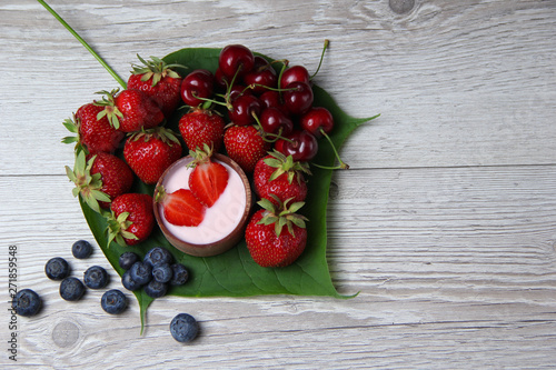 Homemade yogurt with fresh strawberry on a wooden background and some organic strawberries, cherries and blueberries on a big green leaf.