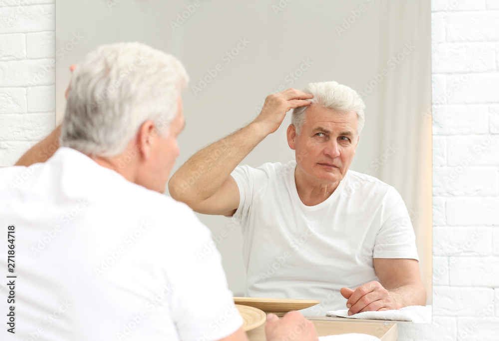 Senior man with hair loss problem looking in mirror indoors