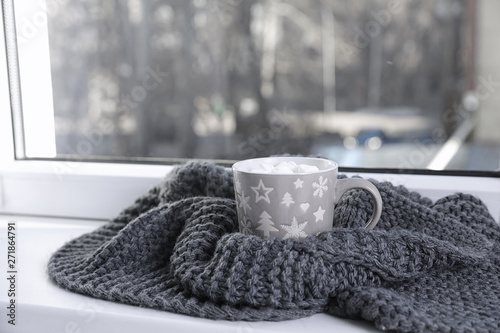 Cup of winter drink and knitted scarf on windowsill. Space for text