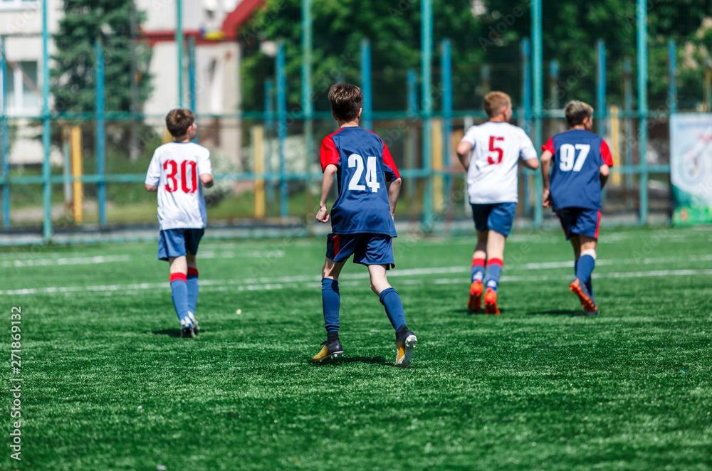 Obraz premium Boys in white and blue sportswear plays football on field, dribbles ball. Young soccer players with ball on green grass. Training, football, active lifestyle for kids concept