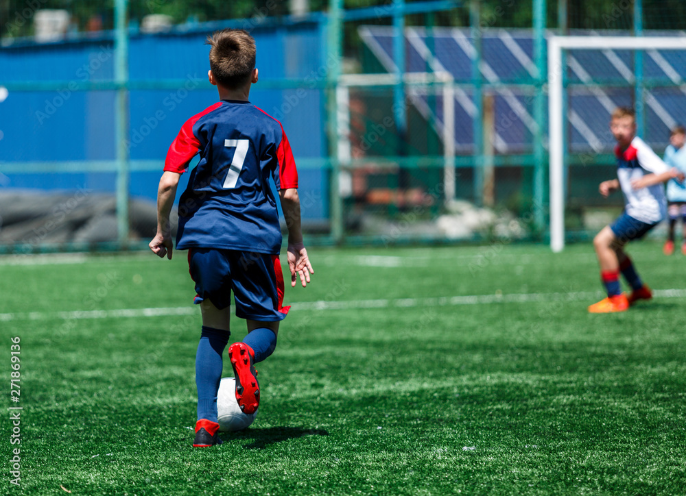 Obraz premium Boys in white and blue sportswear plays football on field, dribbles ball. Young soccer players with ball on green grass. Training, football, active lifestyle for kids concept 