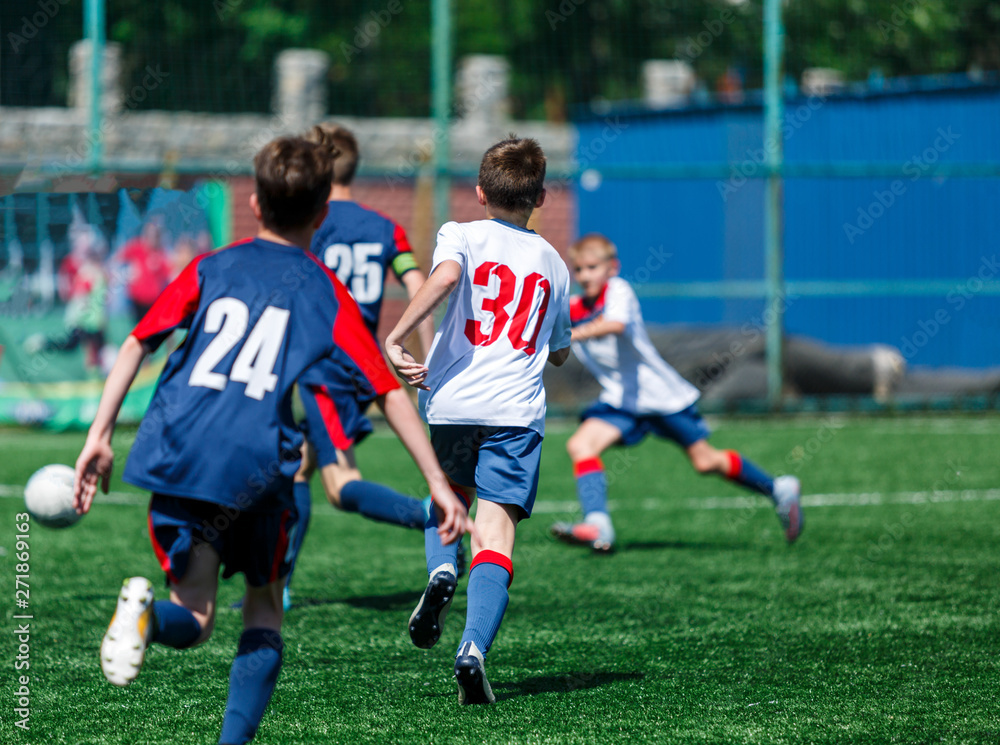 Obraz premium Boys in white and blue sportswear plays football on field, dribbles ball. Young soccer players with ball on green grass. Training, football, active lifestyle for kids concept 