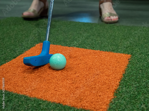 Blue putter on turf next to green golf ball on miniature golf course with woman hitting