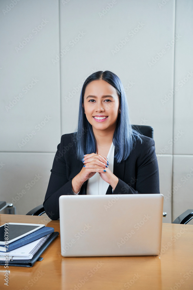 Portrait of pretty young smiling female entrepreneur with blue hair sitting at her office table with laptop and documents Portrait of pretty young smiling female entrepreneur with blue hair sitting at her office table with laptop and documents