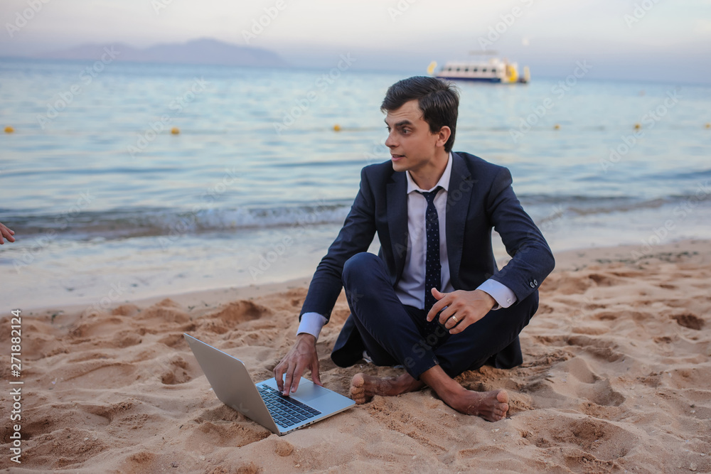lateral view of a young man in suit with laptop sitting on the beach working on the seaside lateral view of a young man in suit with laptop sitting on the beach working on the seaside