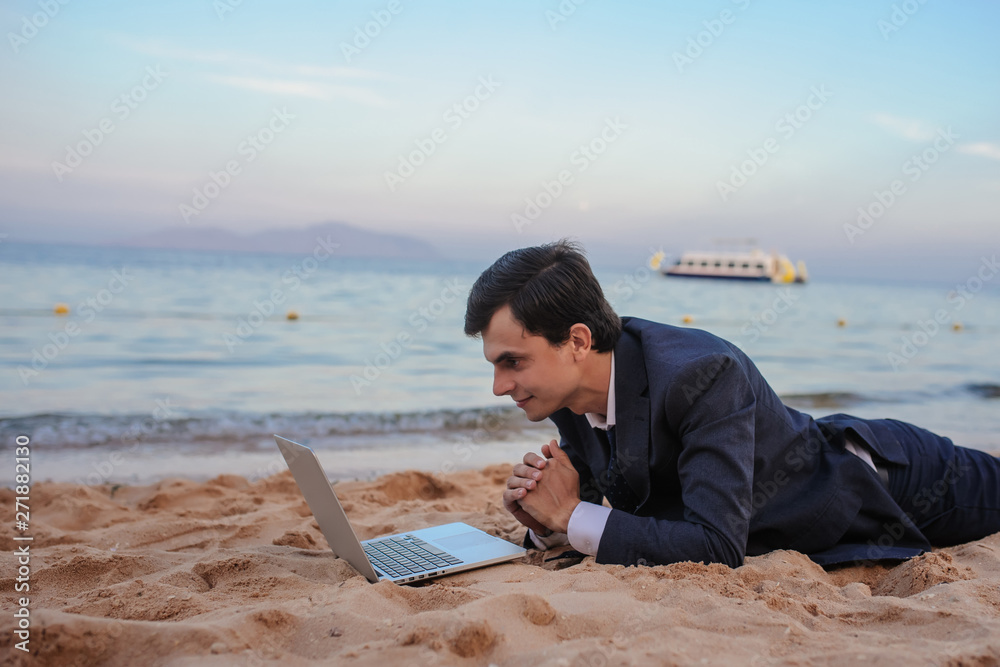 close up photo of a young man in suit with laptop lies on the beach and talking to someone on computer