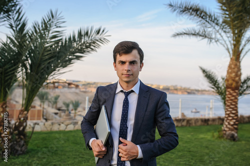 close up photo of a young man in suit with laptop working on green grass near a resort on the seaside photo