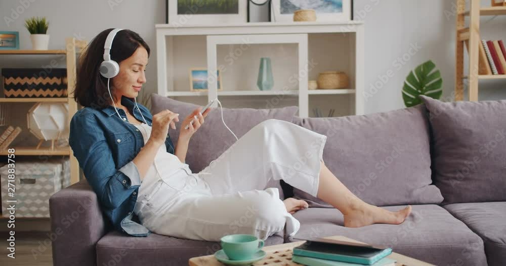 Cheerful girl with dark hair is using smartphone and listening to music in modern headphones sitting on couch in apartment. Lifestyle and youth concept.