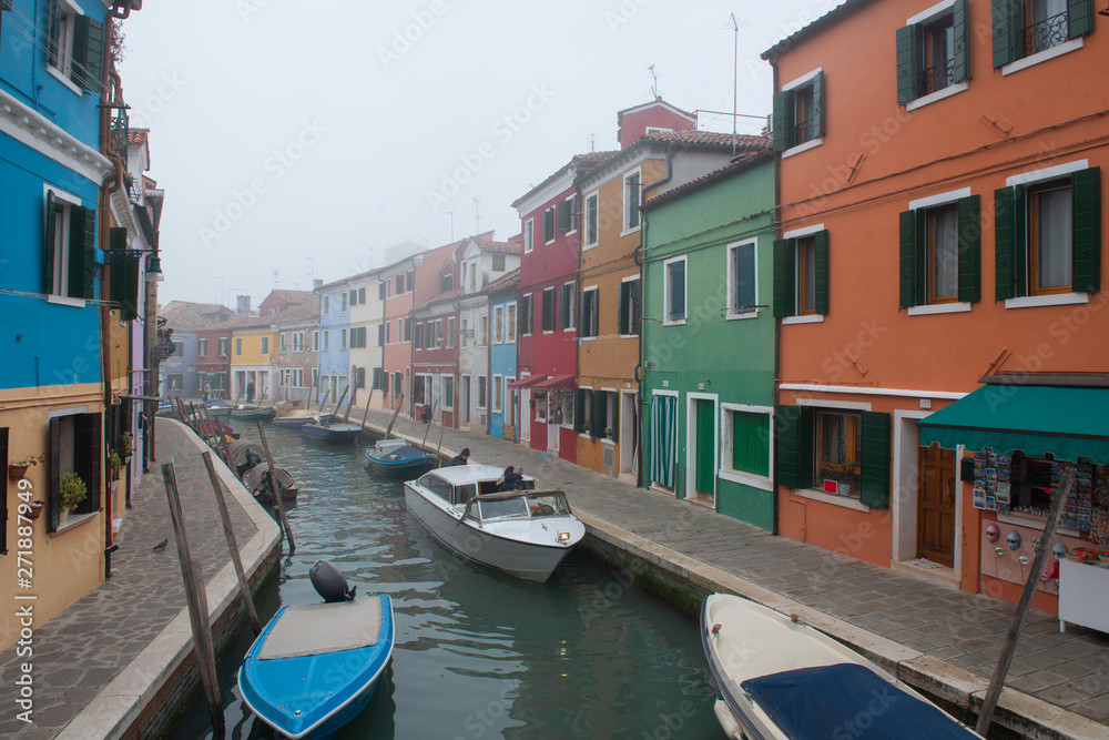 Misty picture of street with colored houses in fog, italian island Burano, province of Venice, Italy, foggy weather. Little beautiful dock with boats, Mediterranean sea.
