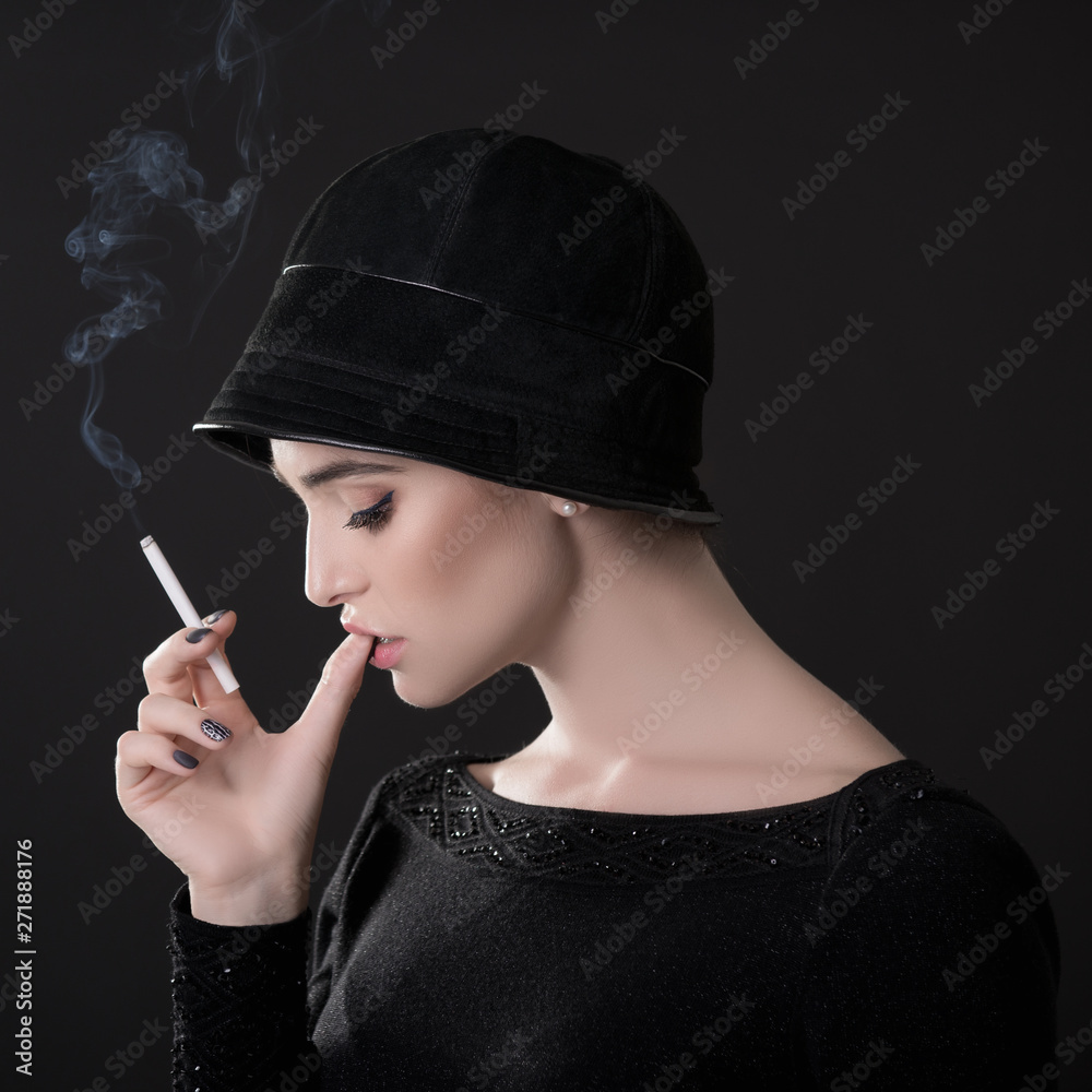 Obraz premium Young fashion woman smoking cigarette in hat and black drees over dark background. Vintage female portrait, styling. Image toned.