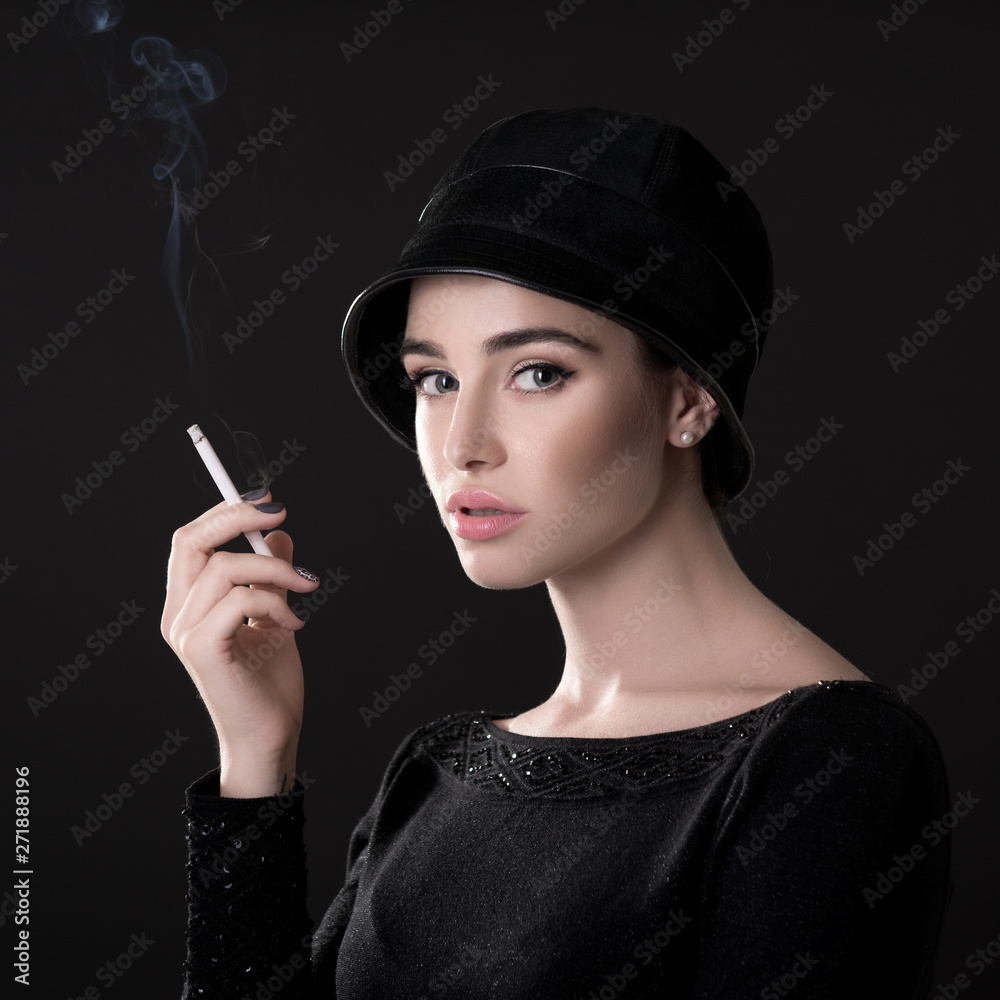 Obraz premium Young fashion woman smoking cigarette in hat and black drees over dark background. Vintage female portrait, styling. Image toned.