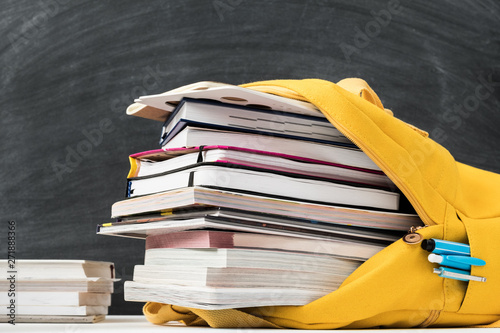 Information overload. Yellow backpack stuffed with books and note pad over empty chalkboard. photo