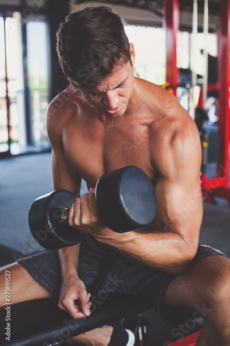 handsome guy with tanned muscular naked torso sits on a bench in the gym and trains biceps with dumbbell