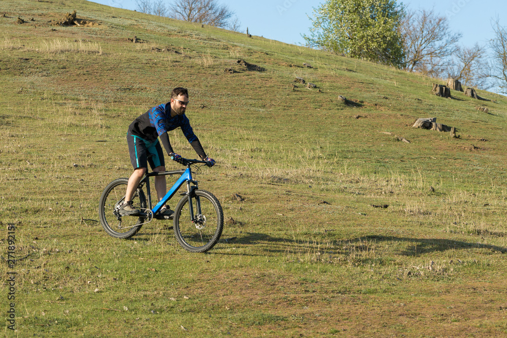 Cyclist in shorts and jersey on a modern carbon hardtail bike with an air suspension fork rides off-road on green hills near the forest Cyclist in shorts and jersey on a modern carbon hardtail bike with an air suspension fork rides off-road on green hills near the forest