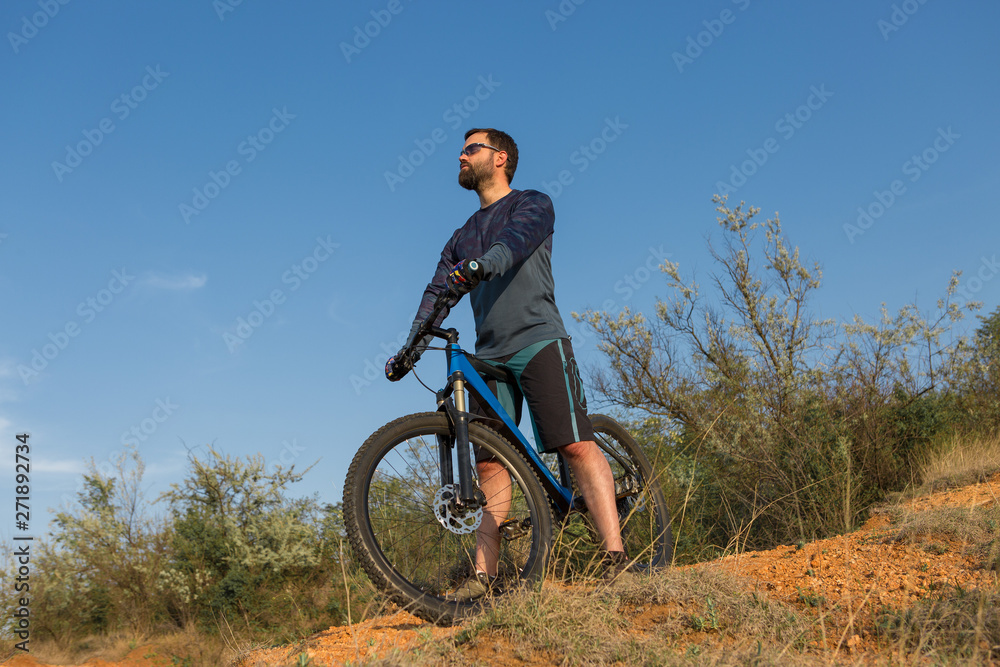 Cyclist in shorts and jersey on a modern carbon hardtail bike with an air suspension fork rides off-road on the orange-red hills at sunset evening in summer Cyclist in shorts and jersey on a modern carbon hardtail bike with an air suspension fork rides off-road on the orange-red hills at sunset evening in summer
