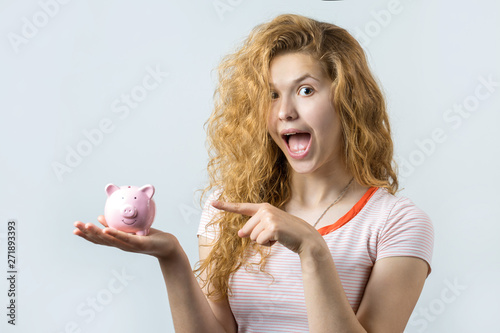 Young red-haired curly girl holding piggy bank and rejoices gesturing. The concept of reliability of cash investments and insurance. 