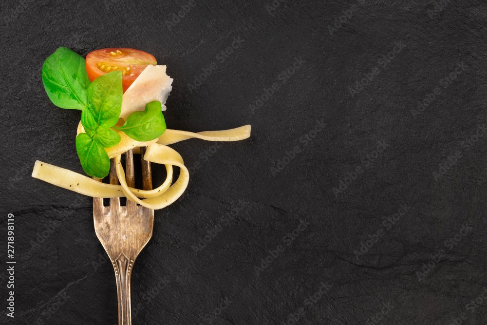 Italian pasta design. A closeup of a fork with pappardelle, basil, Parmesan cheese, tomato, and pepper, shot from above on a dark background with a place for text - obrazy, fototapety, plakaty 