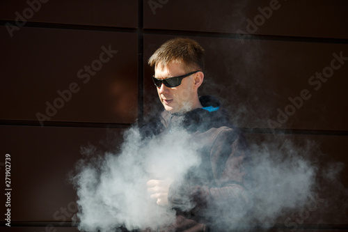 Vape man. An adult bearded man in sunglasses smokes an electronic cigarette on a sunny day outside. Bad habit that is harmful to health. Vaping aclivity.