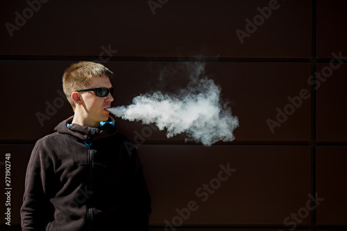 Vape man. An adult bearded man in sunglasses smokes an electronic cigarette on a sunny day outside. Bad habit that is harmful to health. Vaping aclivity.