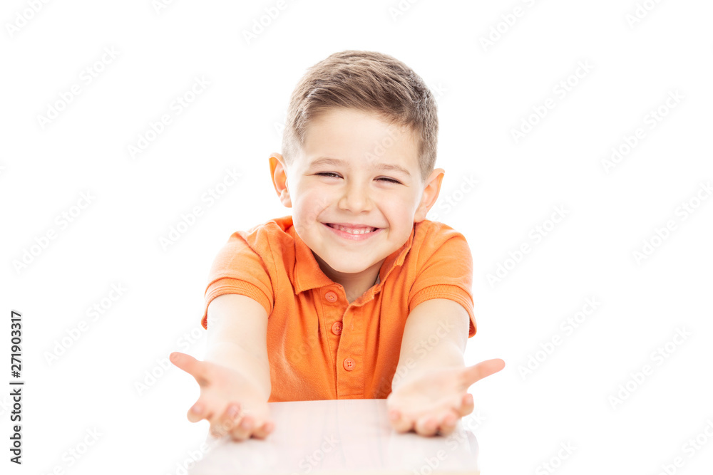 Cute laughing school-age boy in an orange T-shirt sits at the table, arms outstretched. Isolated on a white background. Cute laughing school-age boy in an orange T-shirt sits at the table, arms outstretched. Isolated on a white background.