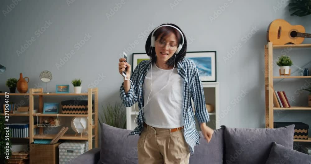 Attractive woman in headphones is dancing listening to music holding smartphone enjoying free time and leisure activity in house. Modern lifestyle concept.