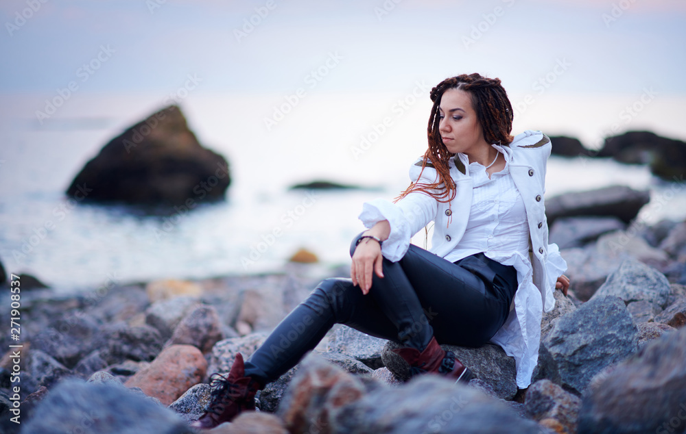 dreadlocks fashionable girl dressed in white jacket and black leather trousers posing near sea in the evening
