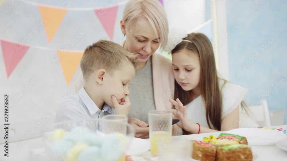 Children are playing the game on smartphone with adult woman sitting by birtyhday table Children are playing the game on smartphone with adult woman sitting by birtyhday table