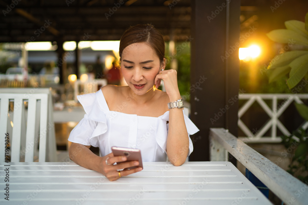 Mixed pretty smiling young woman holding and looking at smart phone on table white outdoor in sunset, enjoying summer vacation travel. in white derss Mixed pretty smiling young woman holding and looking at smart phone on table white outdoor in sunset, enjoying summer vacation travel. in white derss