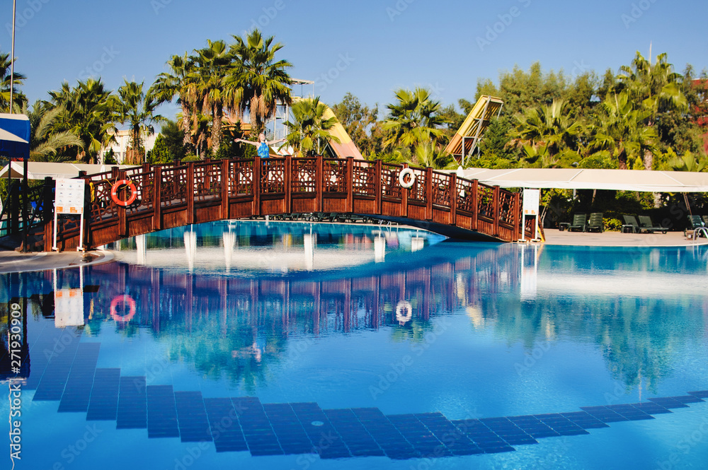 Beautiful young slim girl stands on the bridge through the blue pool. nature, park, outdoors. Turkey. Palms. recreation. Vacation. The path to the house. Sunlight. palm tree