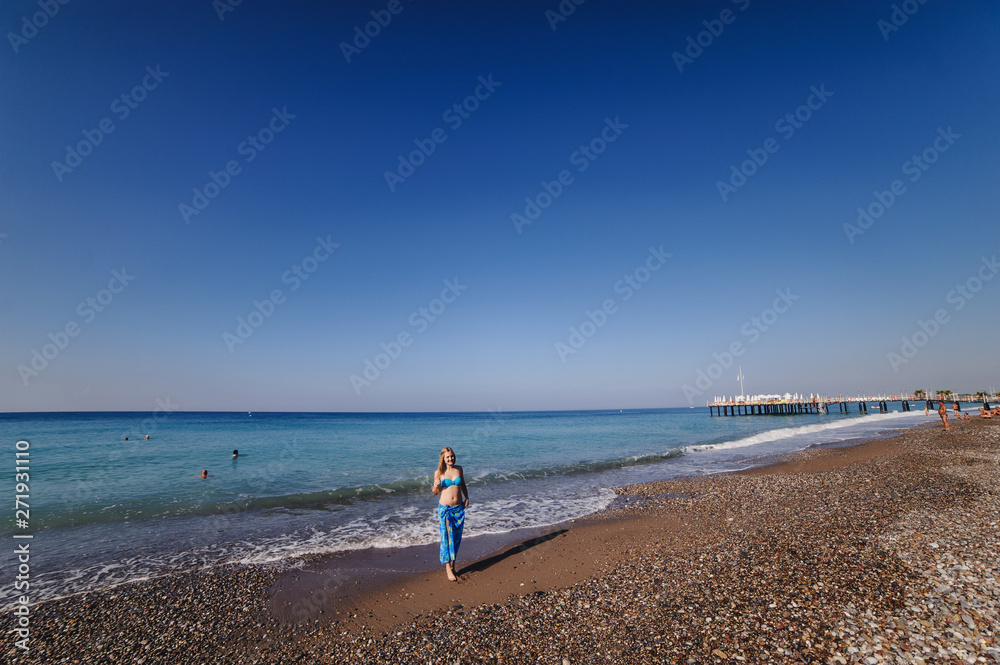 Beautiful young slim girl standing on a sandy beach by the sea. In a blue bathing suit, dress, shawl sarong skirt. Turkey. vacation. Vacation. Sunlight. Place for text Beautiful young slim girl standing on a sandy beach by the sea. In a blue bathing suit, dress, shawl sarong skirt. Turkey. vacation. Vacation. Sunlight. Place for text