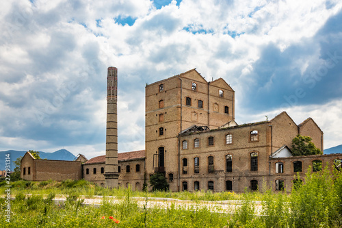 An old disused factory, abandoned and in ruins, with a smashed roof and a chimney. Tall weeds invade the building. Concept of economic bankruptcy. Cloudy sky. Italy, Foligno, Umbria. photo