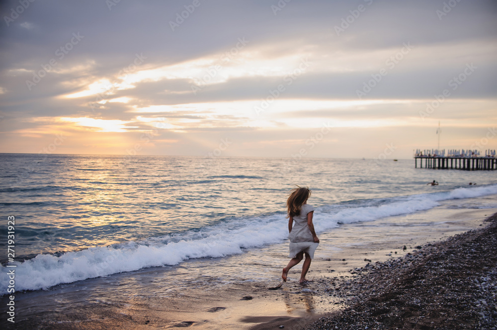 Beautiful young slim girl running on a sandy beach by sea. In a long white dress. Waves. Against the background of the sea. Turkey. Vacation. Sunlight. Place for text. Night. Sunset. horizon. back Beautiful young slim girl running on a sandy beach by sea. In a long white dress. Waves. Against the background of the sea. Turkey. Vacation. Sunlight. Place for text. Night. Sunset. horizon. back