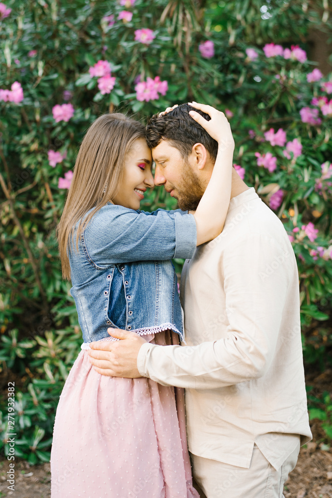 Naklejka premium happy young girl hugs a guy, they rest their foreheads to each other, smile and are happy against the background of spring flowers of pink rhododendron