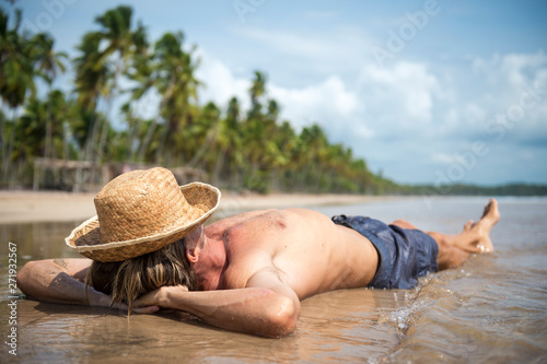 Relaxed tourist wearing a straw sun hat reclining on the shore of a sunny palm-lined tropical island beach 