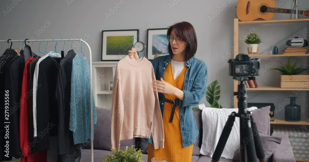 Stylish young woman vlogger is recording video for online blog about trendy clothes holding garment talking and gesturing. Vlogging, people and house concept. Stylish young woman vlogger is recording video for online blog about trendy clothes holding garment talking and gesturing. Vlogging, people and house concept.