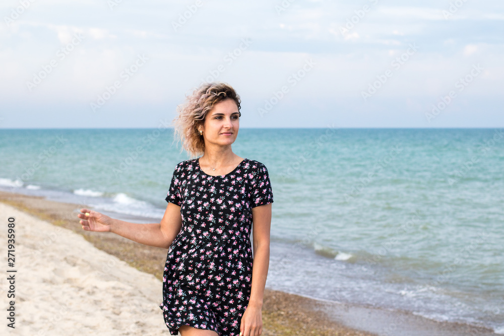 beautiful female with curly hairs in black dress in flowers is standing near water on the beach. Orange sunset light on the face beautiful female with curly hairs in black dress in flowers is standing near water on the beach. Orange sunset light on the face