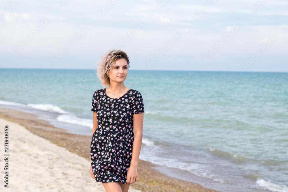 beautiful female with curly hairs in black dress in flowers is standing near water on the beach. Orange sunset light on the face beautiful female with curly hairs in black dress in flowers is standing near water on the beach. Orange sunset light on the face