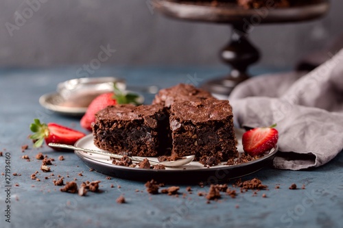pieces of chocolate nut brownie, on saucer with slices of strawberries, crumbs. Blue, grey background.