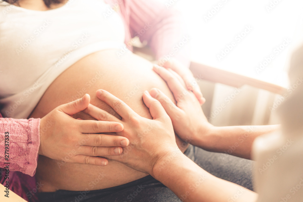 Fototapeta premium Pregnant couple of husband and wife feels love and relax at home. Young expecting woman holds baby in pregnant belly. Father take care of pregnant mother. Concept of maternity and pregnancy care.