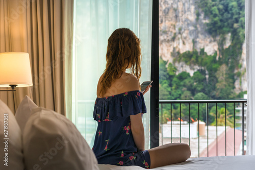 Young woman sits on bed in room of hotel near balcony window and looks in phone display