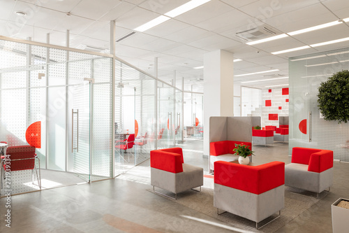 Minsk, Belarus - May 23, 2019: Corner of modern office with white walls, gray floor, open space area with red-white armchairs and rooms behind glass wall photo