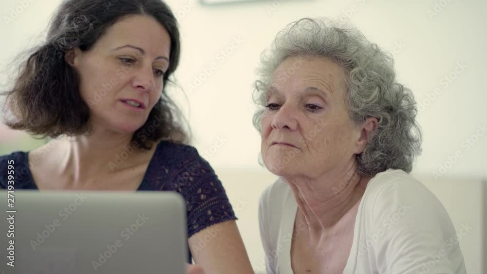 Two smiling women using laptop at home. Happy beautiful ladies sitting at table with modern laptop and talking. Technology concept Two smiling women using laptop at home. Happy beautiful ladies sitting at table with modern laptop and talking. Technology concept